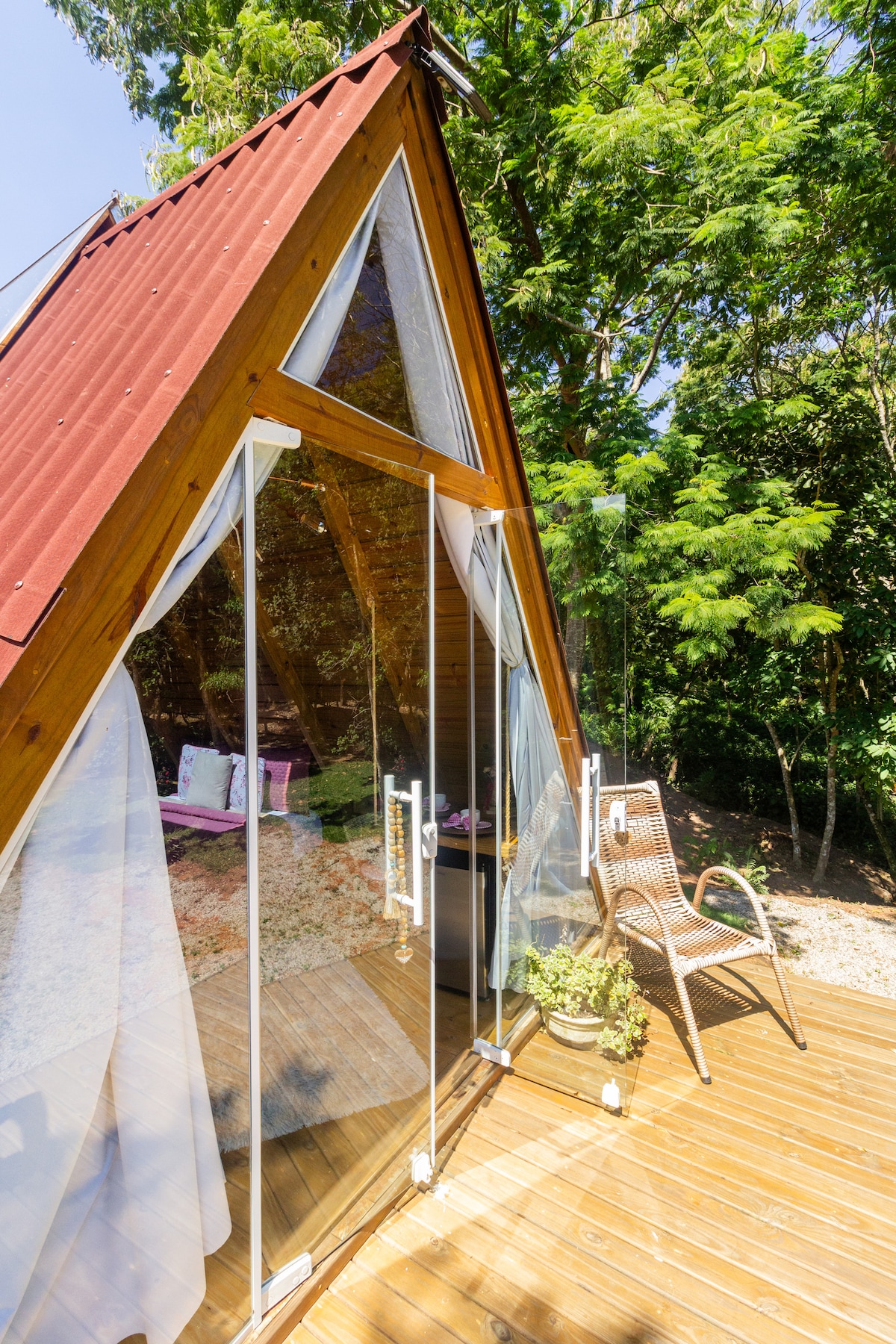 An A-frame structure is showcased with large glass doors that open to a wooden deck. Bright natural light filters through the glass, illuminating the area. A comfortable chair is positioned on the deck, accompanied by greenery and tree foliage in the background.