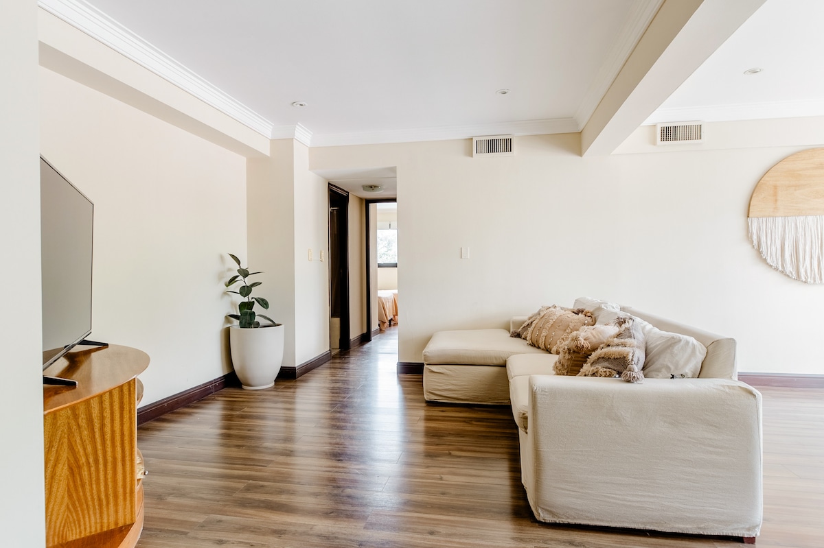 A spacious living area features a light-colored sectional sofa positioned near a television. A decorative round wall piece adds interest to the neutral walls. A potted plant sits on the floor, contributing to a sense of openness in the well-lit room.