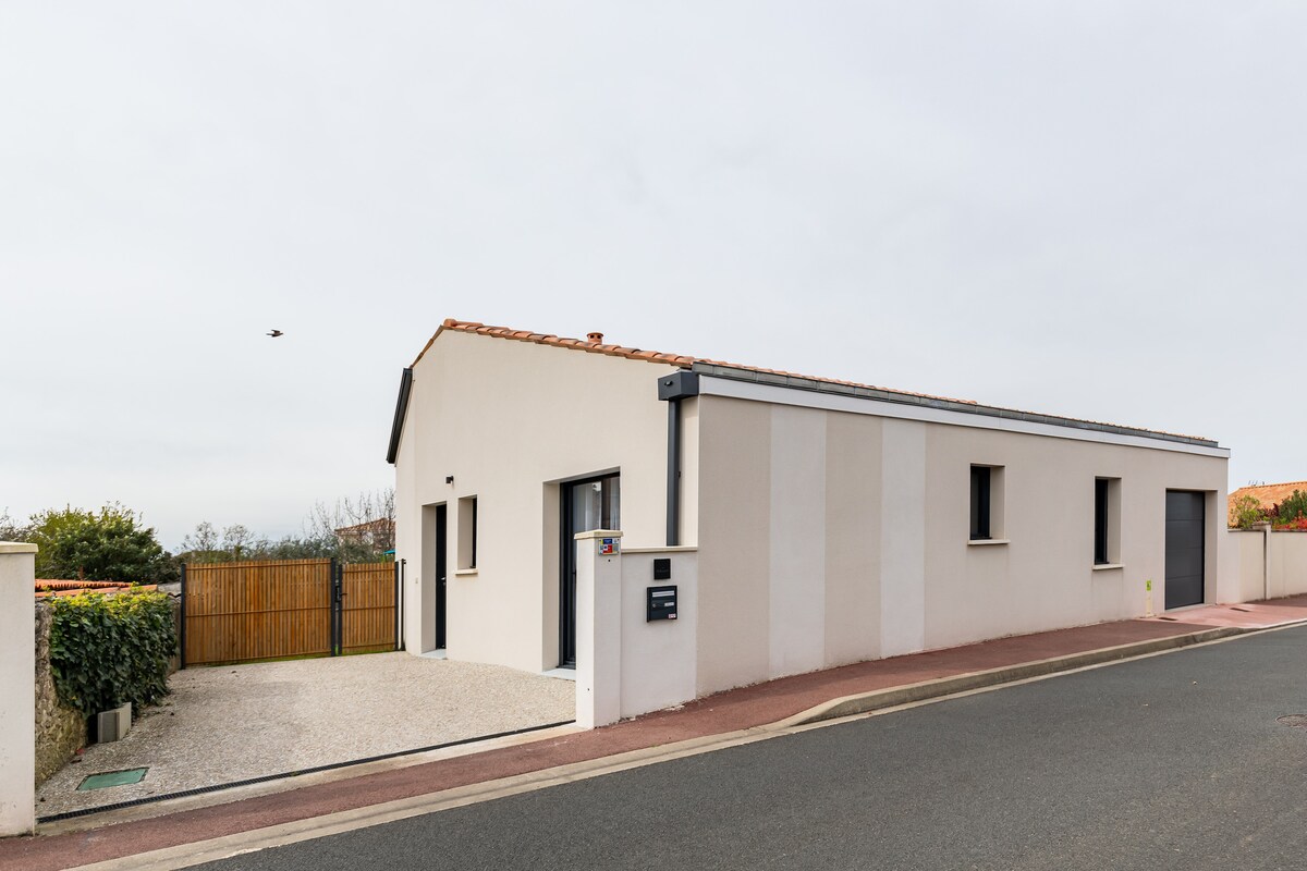 A modern single-story house is shown from the street, featuring a simple exterior design with horizontal stripes. A gravel pathway leads to the entrance, bordered by low fencing and greenery. The surrounding area is quiet, with minimal traffic visible on the road.