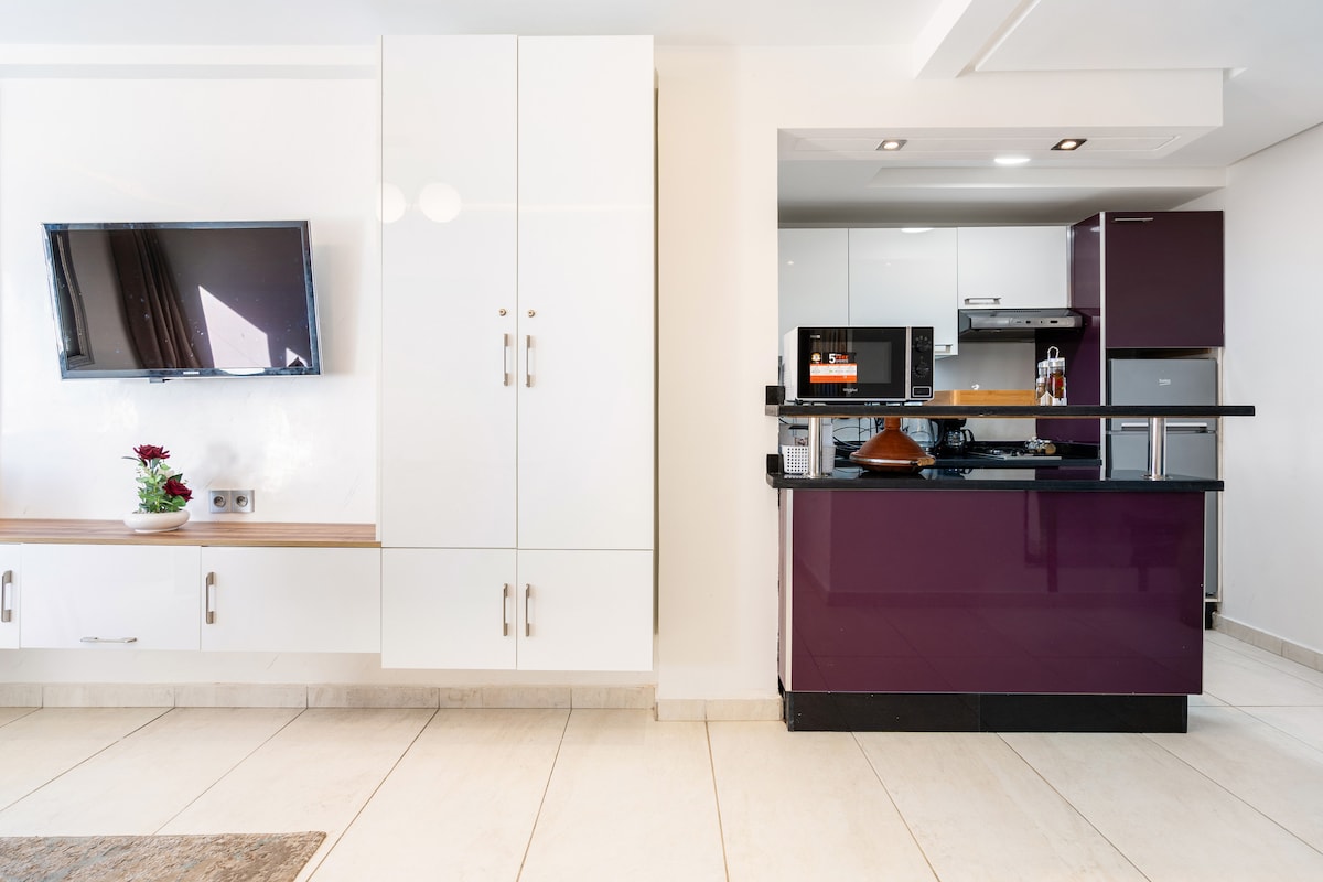A modern kitchen area is visible, featuring sleek purple cabinets complemented by white cabinetry. A coat of natural light fills the space, highlighting a mounted television and a small decorative plant on a lower shelf. Stainless steel appliances are arranged efficiently, enhancing functionality.