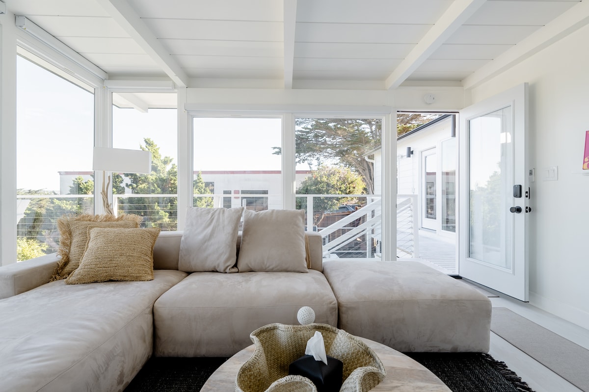 A spacious living area features a light-colored sectional sofa adorned with textured cushions. Natural light floods in through floor-to-ceiling windows, illuminating the minimalist decor. A round coffee table with a decorative bowl sits centered in front of the seating area, enhancing the serene atmosphere.