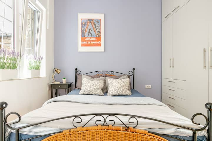 Camera da letto luminosa e tranquilla con un accogliente letto matrimoniale, lenzuola morbide e molta luce naturale. Presenta un elegante telaio del letto in ferro, un armadio a muro, arredi color lavanda e un poster retrò di James Bond, perfetto per un soggiorno riposante nel centro di Belgrado.