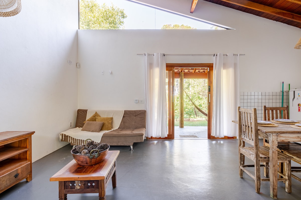 A spacious living area is presented with a large window allowing natural light to fill the space. A comfortable sofa is positioned beside a rustic wooden coffee table, while a wooden dining table for four is visible. Light curtains flutter gently by the entrance, which opens to a green outdoor view.