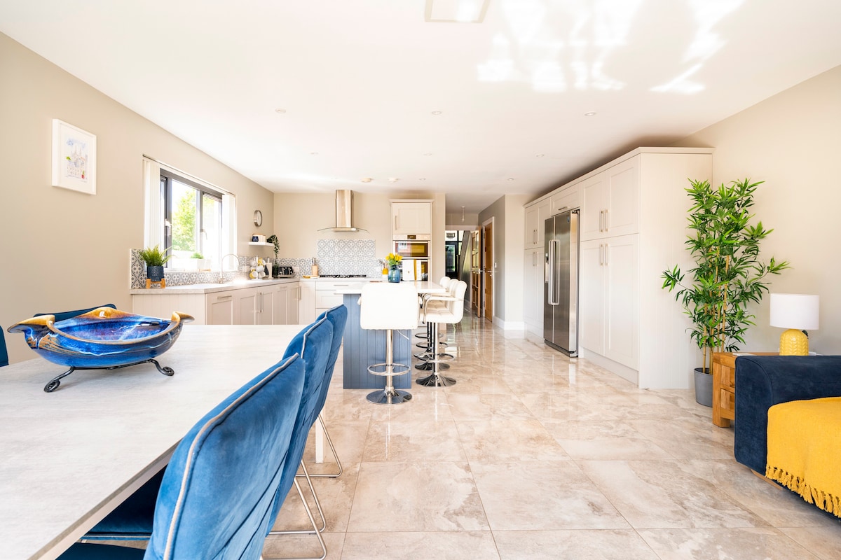 A spacious kitchen and dining area is depicted, showcasing modern cabinets and an island countertop. A large dining table with blue chairs is positioned alongside, while sleek appliances and ample natural light create a functional atmosphere. Decorative plants add a touch of vibrancy to the space.