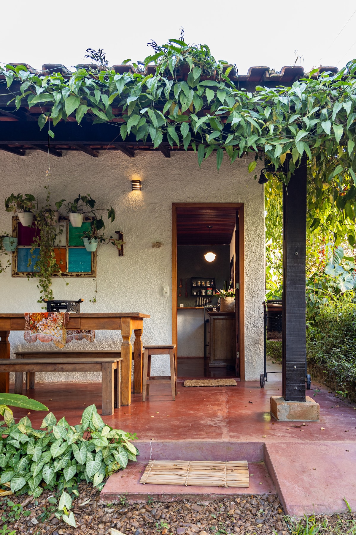 A charming outdoor seating area features a wooden table and benches beneath a covered porch. Green vines hang gracefully from the roof, while potted plants add a touch of nature. The entrance leads to an inviting interior, visible through an open door.