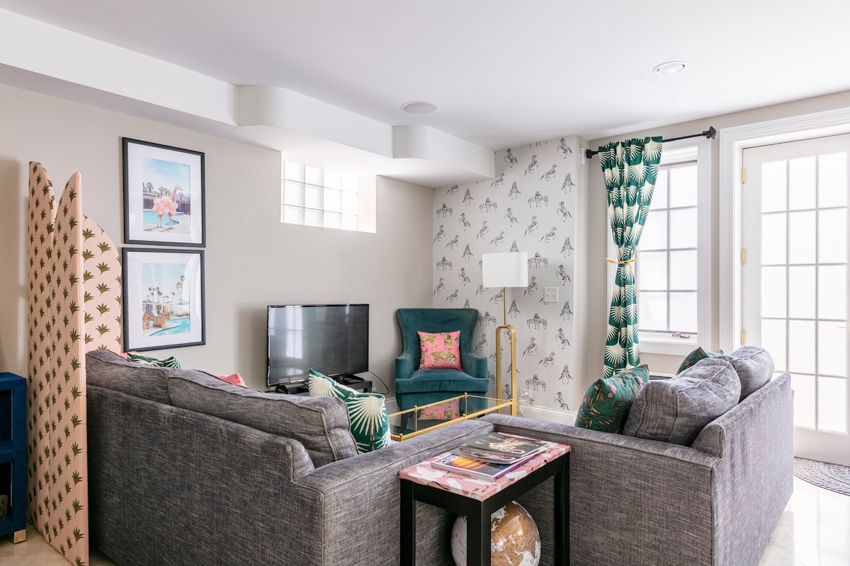 A cozy living room features a gray sectional sofa and a teal accent chair, complemented by patterned curtains. A TV is positioned on a low stand, with framed artwork on the walls. Natural light filters through multiple windows, enhancing the vibrant decor.