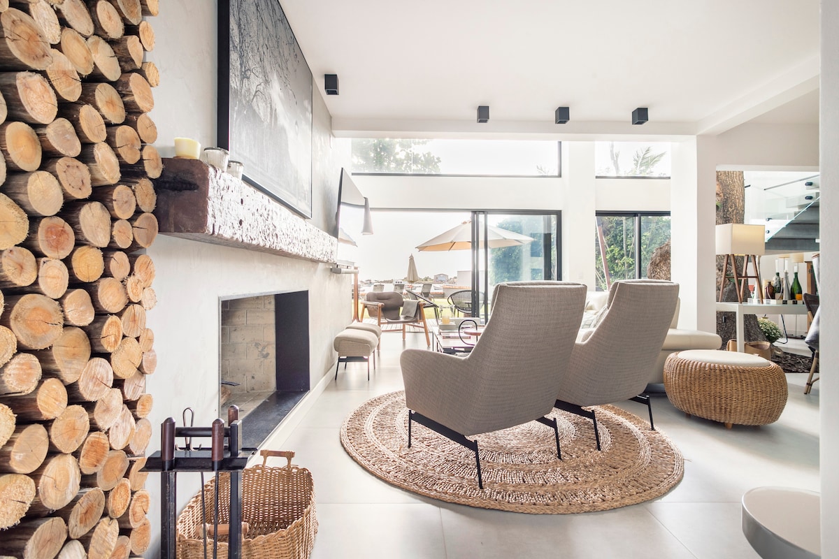 A bright living area features contemporary furnishings, including two armchairs positioned around a natural fiber rug. A stack of wooden logs creates a rustic accent beside a sleek fireplace. Large windows provide views of the outdoors, allowing natural light to fill the space.