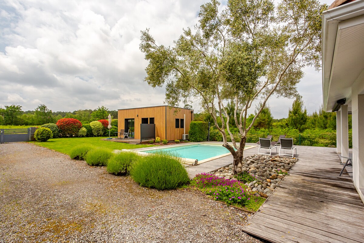 A spacious outdoor area features a refreshing pool surrounded by neatly trimmed grass and colorful flower beds. A wooden structure is visible in the background, along with lounge chairs placed beside the pool. Tall trees and shrubs provide a natural border, enhancing the tranquil environment.
