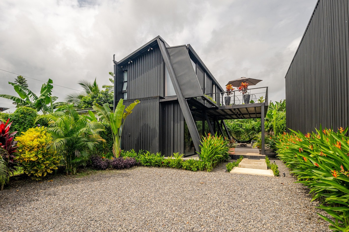 A modern structure with a distinctive angular design is surrounded by lush greenery and vibrant tropical plants. A gravel pathway leads to the entrance, while an elevated balcony with dining furniture overlooks the landscape, providing a space for outdoor relaxation.