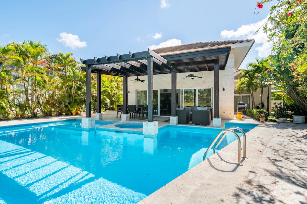 A private villa features a spacious outdoor area with a large swimming pool surrounded by palm trees and lush greenery. A covered terrace with seating and dining options overlooks the pool, providing a serene atmosphere for relaxation.
