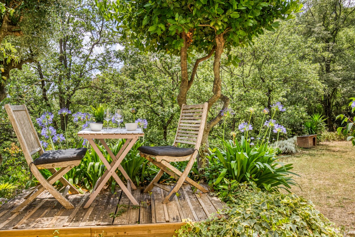 A small wooden deck features a table set with glassware and white dishware for dining. Two wooden chairs are positioned around the table, surrounded by lush greenery and blooming flowers, creating a serene outdoor dining space.