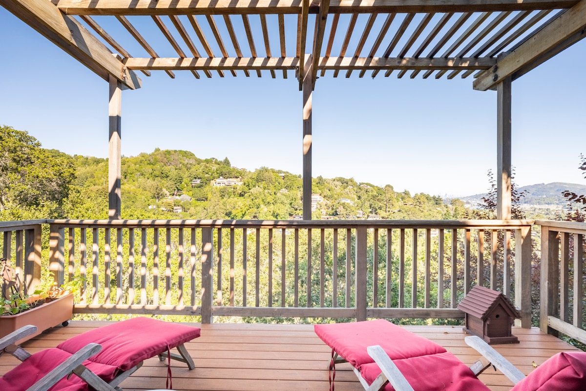 A sun-drenched deck is framed by wooden beams, offering expansive views of the surrounding greenery. Two comfortable lounge chairs with red cushions are positioned for relaxation. A wooden railing encircles the space, enhancing the outdoor ambiance.