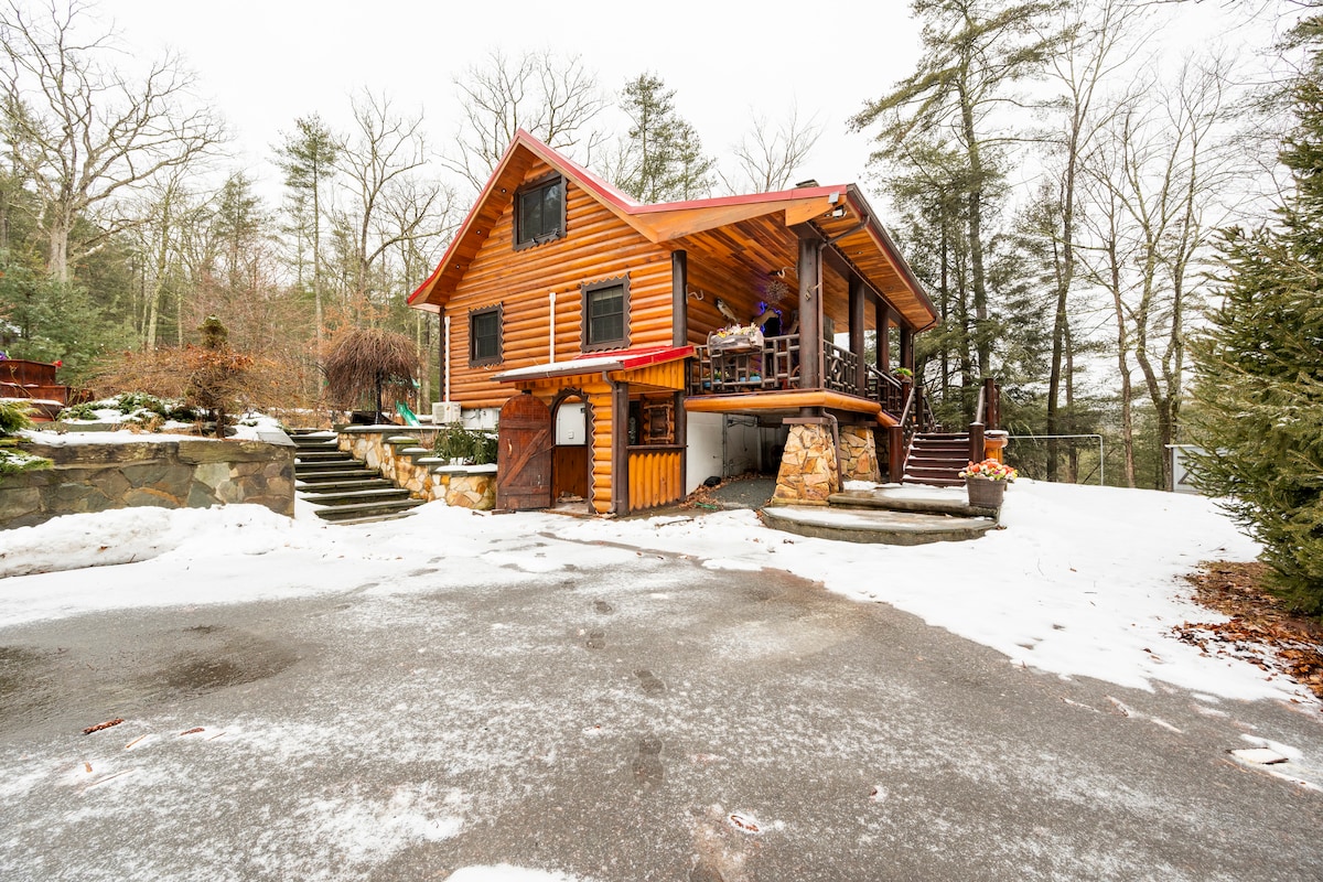A multi-level wooden house is seen surrounded by a snowy landscape. The structure features a distinctive peaked roof and large windows. Steps lead up to the entrance, while a stone wall and a range of trees frame the property, contributing to a secluded ambience.