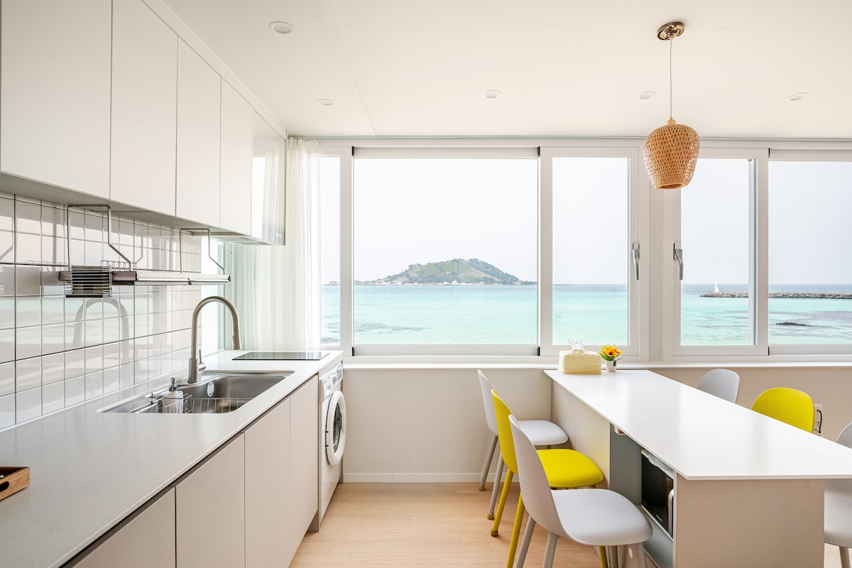 A bright kitchen space is characterized by sleek cabinetry and a large window overlooking a serene view of the ocean and nearby island. A dining table is set with chairs featuring yellow accents, enhancing the light ambience of the room.