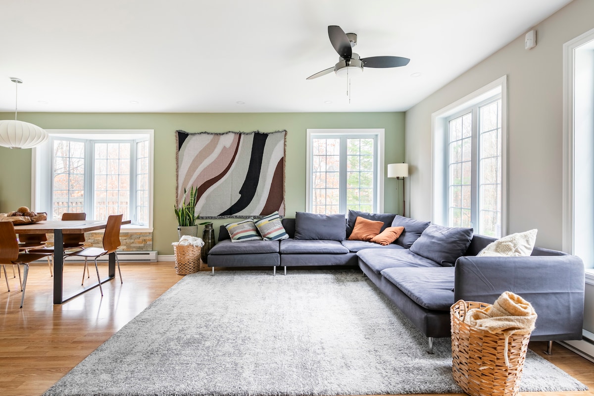 An open-concept living space features a sectional sofa adorned with cushions, adjacent to a dining area with a round table and several chairs. Large windows allow natural light to enhance the light-colored walls, while an artistic wall hanging adds a pop of color to the serene environment.