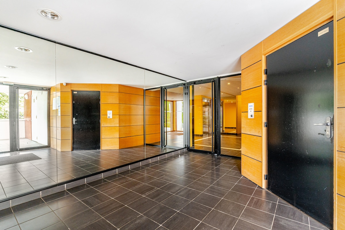 A welcoming lobby area showcases sleek, modern design elements with wooden paneling and expansive mirrors. The space features dark tile flooring and glass doors that provide access to the outdoors, emphasizing a bright and spacious entryway.