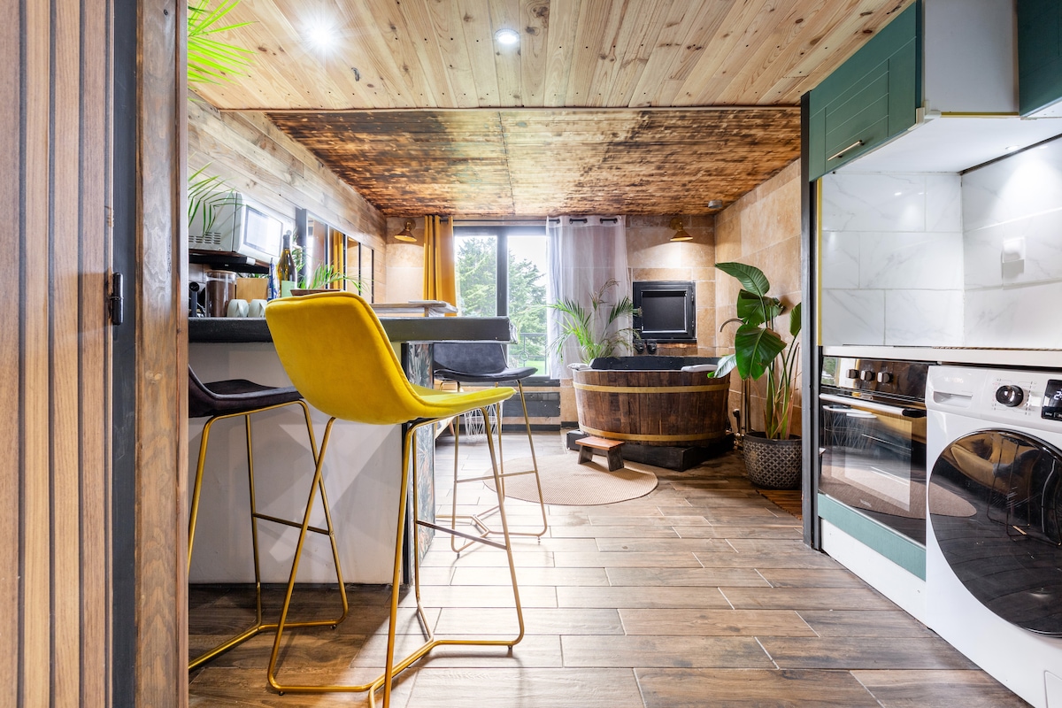 An open-plan living space is featured, showcasing a modern kitchen and dining area. A spacious bathtub is positioned near a window, with green plants adorning the space. Natural wood ceilings add warmth, while yellow bar stools provide a pop of color.