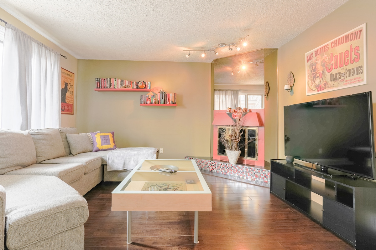 A cozy living area is presented, featuring a spacious sectional couch and a glass coffee table. A television sits on a dark stand, while vibrant wall art and a bookshelf filled with books add character. Natural light filters through sheer curtains.