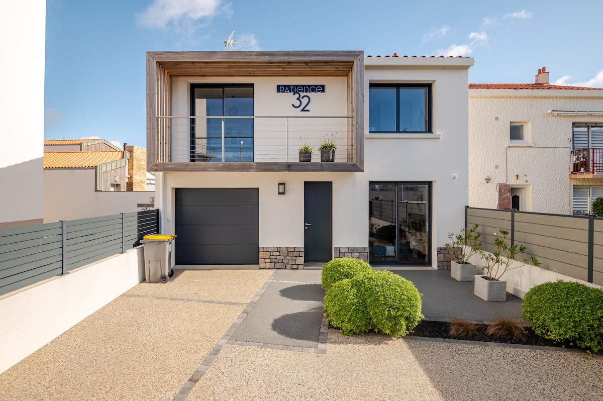 A modern, two-story house features a wooden balcony above the entrance, which is adorned with greenery. A dark garage door is positioned on one side, while neatly trimmed shrubs and planters line the pathway leading up to the front door. Clear blue skies are visible overhead.