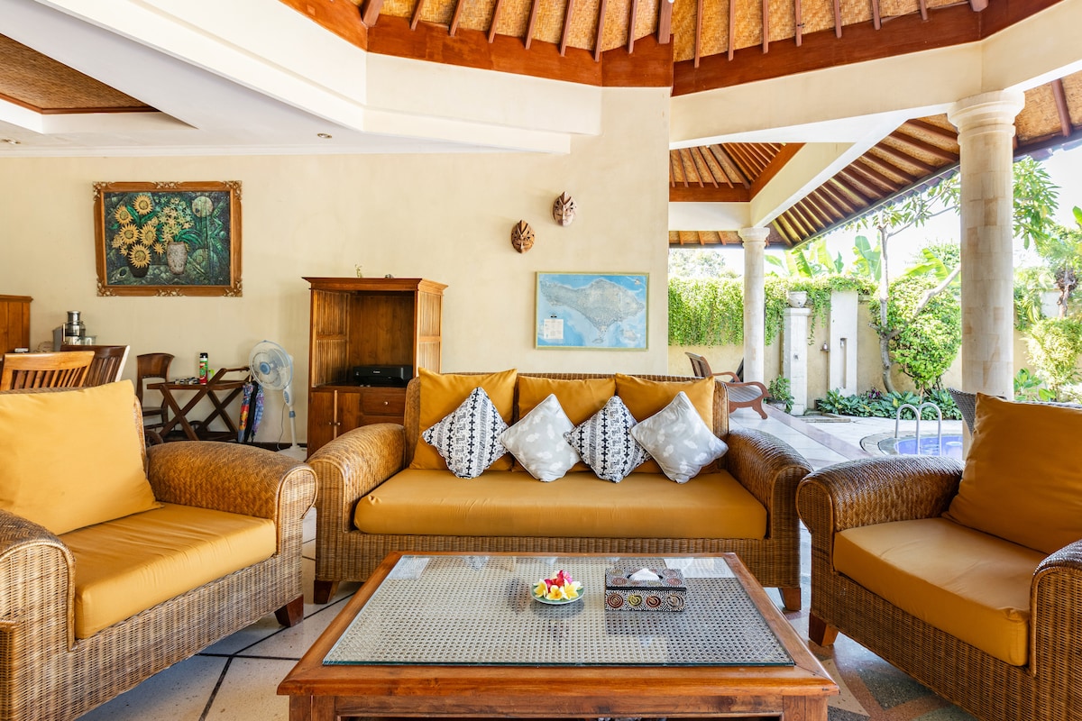 A spacious lounge area features comfortable woven seating with golden cushions positioned around a wooden coffee table. Natural light floods the space through the surrounding open design, highlighting the tropical ambiance and subtle artwork adorning the walls.