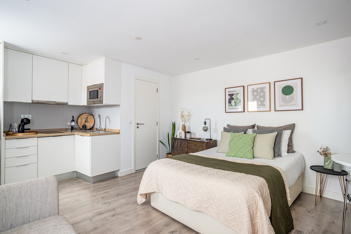 A studio apartment interior features a spacious bed adorned with neutral-colored linens. A kitchenette is visible, equipped with modern appliances including a microwave. Artwork frames are arranged on the wall above the bed, contributing to a tidy and inviting ambiance.