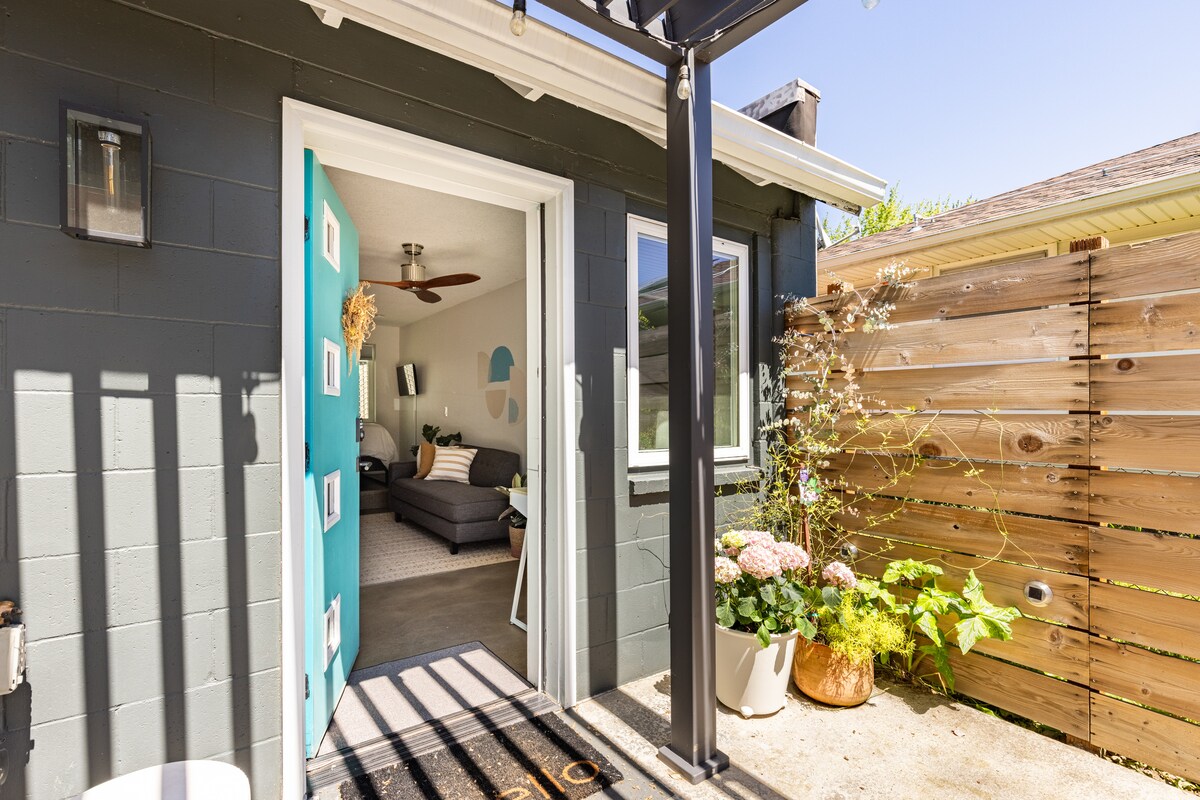The entrance to the studio apartment is framed by a wooden pergola, showcasing a blue door with a modern design. Potted plants and flowers enhance the outdoor space, while a cozy seating area is visible in the background through large windows.