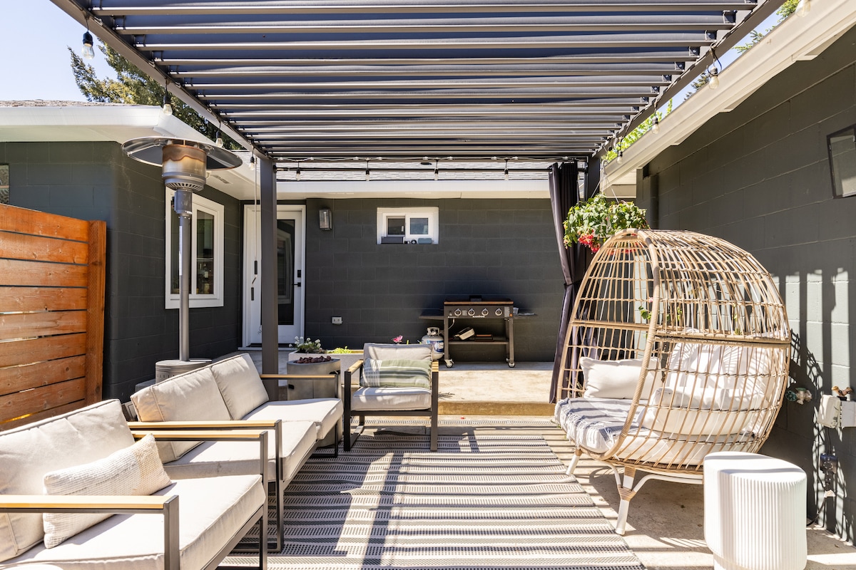 An outdoor seating area is provided under a sleek pergola, featuring a mix of comfortable couches and a woven hanging chair. A subtle brown and white striped rug lays beneath, enhancing the relaxed ambiance. Potted plants and decorations add greenery to the space.