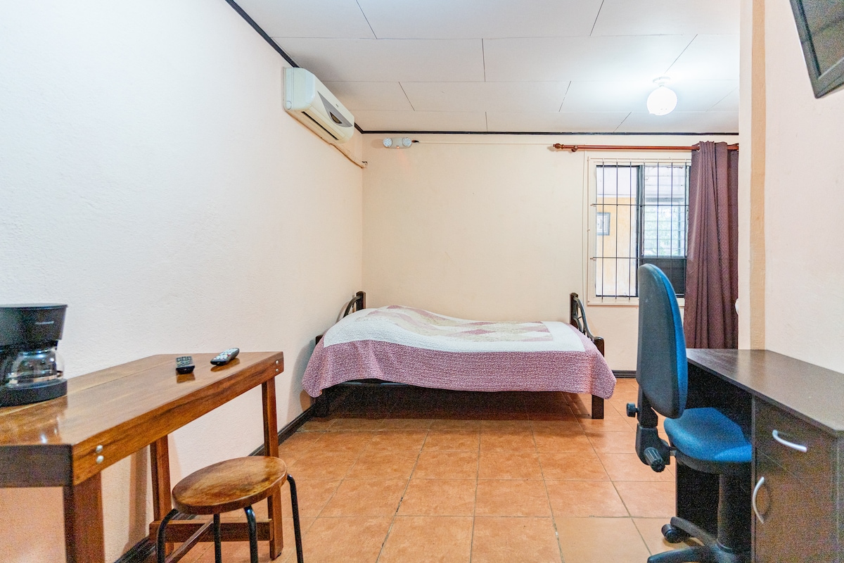 A simple room is featured with a spacious bed covered by a light-colored blanket. A wooden table and a stool are positioned near a window, while a desk and chair occupy the opposite corner. A wall-mounted air conditioner and a ceiling light provide comfort and illumination.