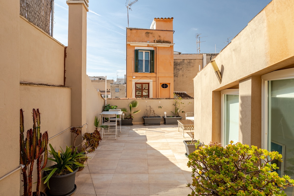 A spacious terrace is displayed, featuring multiple potted plants and a dining table set for outdoor enjoyment. The surrounding walls provide privacy, while the bright sky and nearby buildings create an inviting atmosphere. Natural stone flooring enhances the aesthetic of this outdoor space.