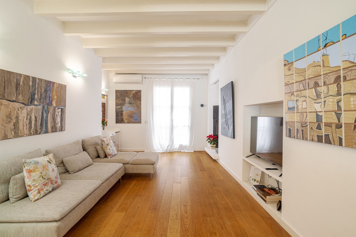A spacious living area features a comfortable L-shaped sofa accompanied by decorative cushions. Natural light filters through sheer curtains, illuminating the wooden floor. Artwork hangs on the walls, adding character to the room, while a television is mounted for viewing convenience.
