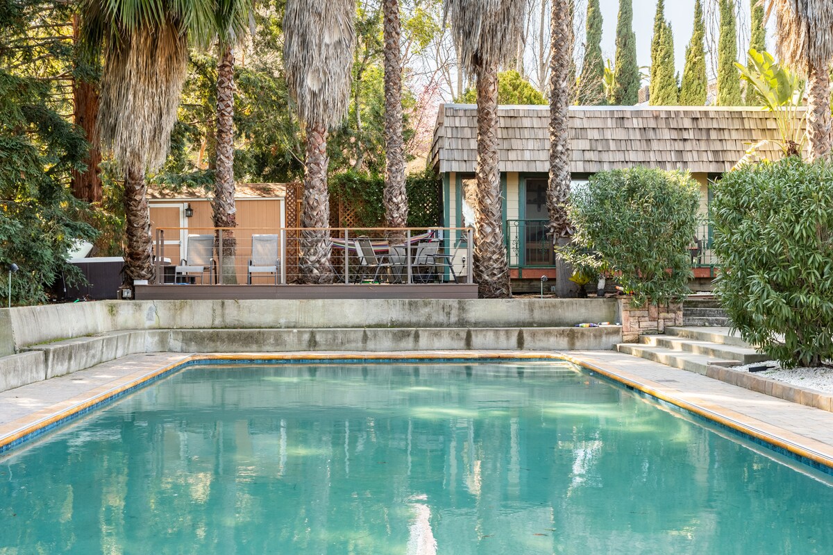 A salt-water pool is visible in the foreground, surrounded by tall palm trees and a landscaped area. In the background, a guest house with a wooden roof and outdoor seating area is nestled among greenery, creating a serene setting.