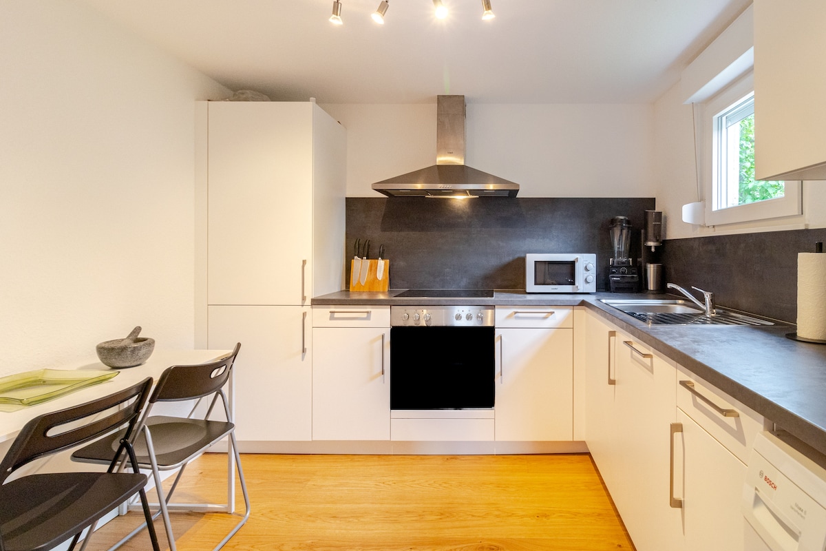 A modern kitchen is equipped with sleek white cabinetry and a dark countertop. Stainless steel appliances, including an oven and microwave, are visible. A small dining area features two black folding chairs and a simple green placemat, while natural light streams in from a nearby window.