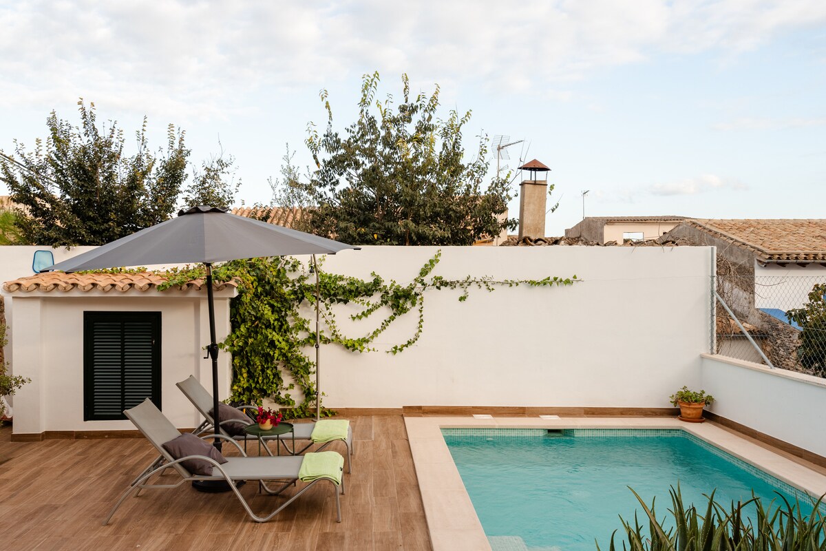 A private pool area is presented, featuring a turquoise pool bordered by wooden decking. Two loungers with green cushions are positioned under a gray umbrella, surrounded by lush greenery and trees. A white wall with climbing plants creates a serene atmosphere for relaxation.