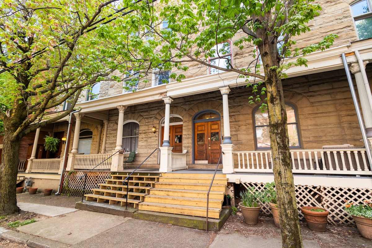 A historic building features a welcoming entrance with double wooden doors framed by large windows. A set of stairs leads up to a covered porch, while lush green trees provide shade. Potted plants enhance the inviting exterior, showcasing the charm of the neighborhood.