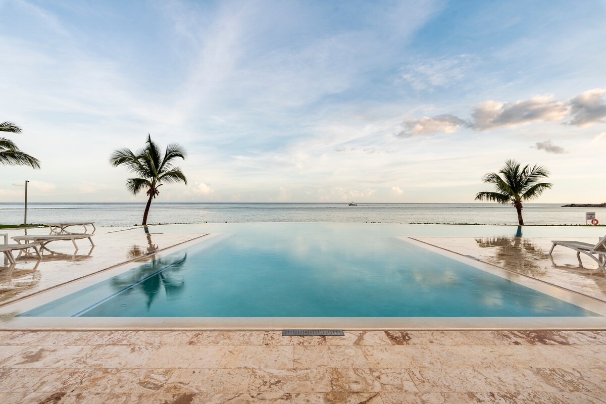 An expansive infinity pool reflects the sky, seamlessly connecting with the ocean horizon. Flanking the pool are palm trees, gently swaying in the breeze. The surrounding area features light-colored stone flooring and lounge chairs, inviting relaxation by the water's edge.