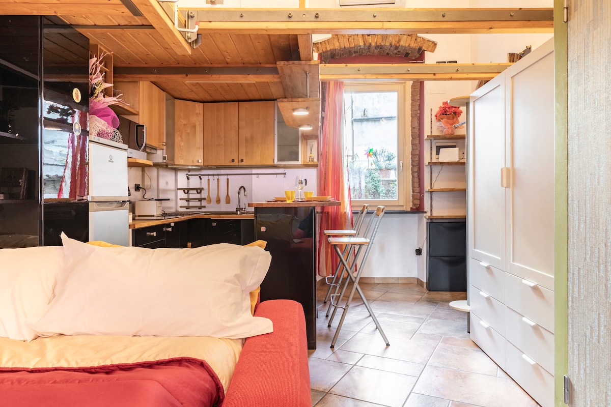 An open-plan living area features a comfortable bed adjacent to a modern kitchen equipped with essential appliances. A dining space with two high stools enhances the layout. Natural light filters through a window adorned with sheer curtains, creating a welcoming ambiance.