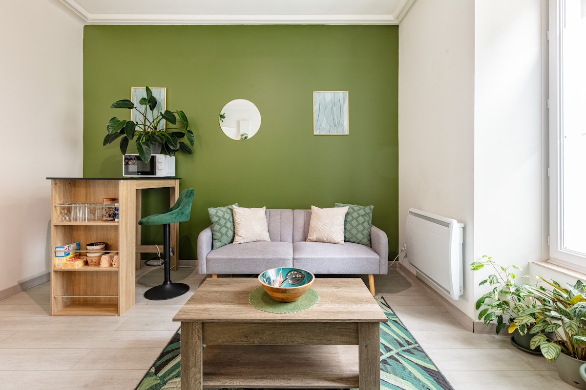 A comfortable living area features a light gray sofa with decorative cushions, complemented by a wooden coffee table. A small bar area with shelves holds various items, while a green accent wall adds a fresh touch. Houseplants introduce natural elements near the window, enhancing the space.
