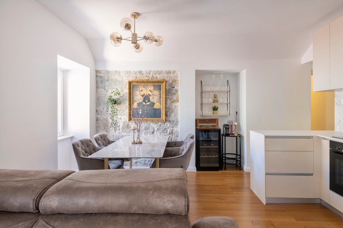 A dining area features a sleek table surrounded by comfortable chairs, illuminated by a stylish chandelier. Light floods in through large windows, highlighting a unique artwork on the wall. A minimalist kitchen space is showcased nearby, with modern appliances and storage options.