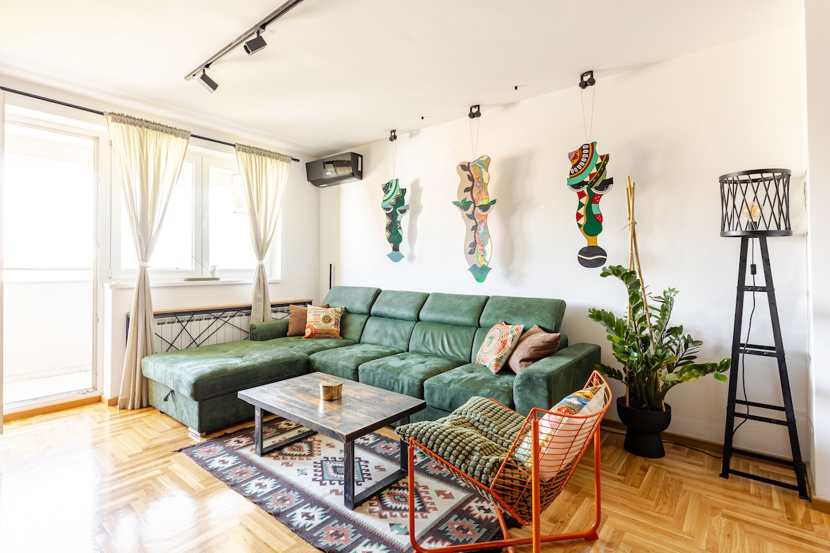 A spacious living area features a green sectional sofa complemented by patterned cushions. Colorful wall art adds character, while a low coffee table is positioned centrally. Natural light pours in through large windows, and a small indoor plant brings a touch of greenery to the space.