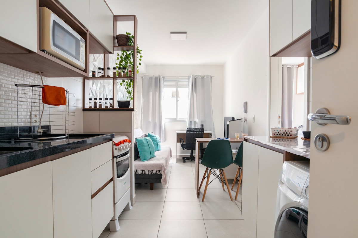 The open-plan living space features a modern kitchen with sleek cabinetry and a countertop. A comfortable sofa bed and a small dining table are visible, adjacent to a window with natural light. Green plants add a touch of vibrancy to the decor.