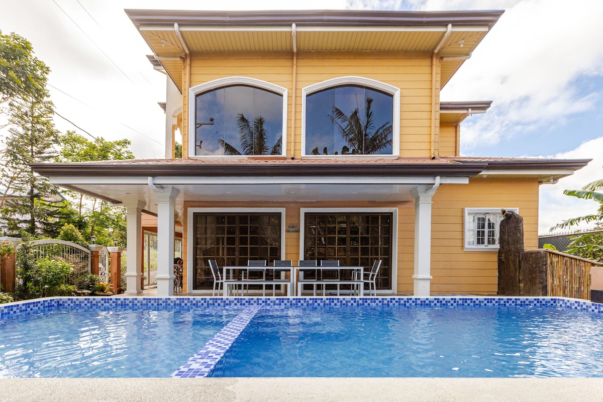 The exterior of a two-story house is showcased, featuring large arched windows that reflect the surroundings. A private pool is seen in the foreground, with a blue mosaic tile border. The outdoor dining area is visible, furnished with a table and chairs beside the pool.