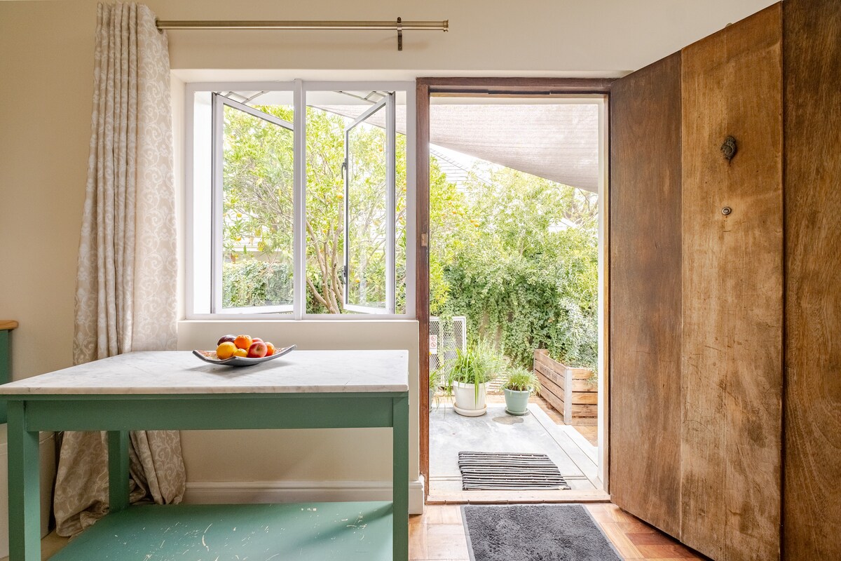 A dining area features a light-colored table with a bowl of fresh fruit. Large windows allow natural light to fill the space, while an attractive wooden door opens to a lush garden area, enhancing the indoor-outdoor connection.