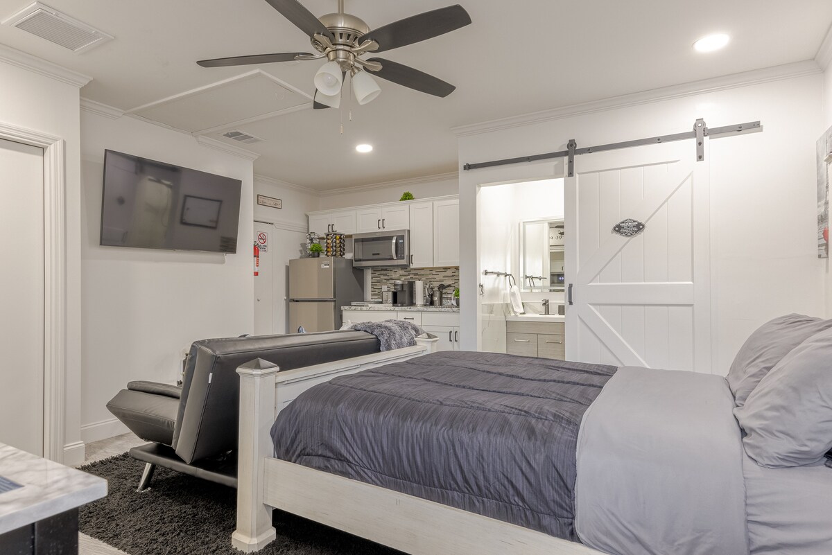 A well-appointed suite features a spacious bed with dark bedding, a comfortable seating area with a grey lounge chair, and a flat-screen television mounted on the wall. A kitchenette with modern appliances is visible in the background, complemented by white cabinetry and a light-colored countertop.