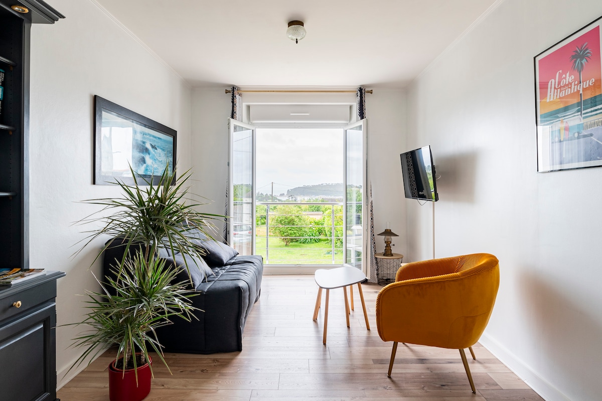 A bright living area features large open doors leading to a scenic view. A comfortable black sofa and a vibrant orange armchair are positioned around a small wooden table. A potted plant adds a touch of greenery, while a television is mounted on the wall.