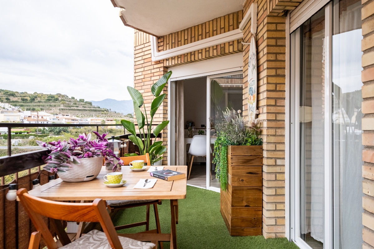 An inviting balcony features a wooden table set with two bowls and a folded napkin. Surrounding plants add greenery, while a wooden planter sits against the brick wall. Large sliding doors provide access to the indoor living area, enhancing the connection between spaces.