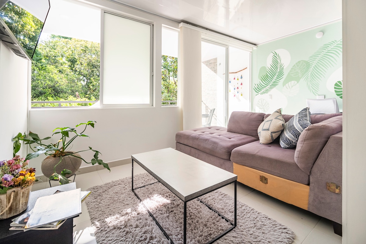 A cozy living area is shown, featuring a spacious sectional sofa adorned with patterned pillows. A light wood coffee table sits on a soft rug, while large windows allow natural light to fill the space. Green plants add a touch of nature to the environment.