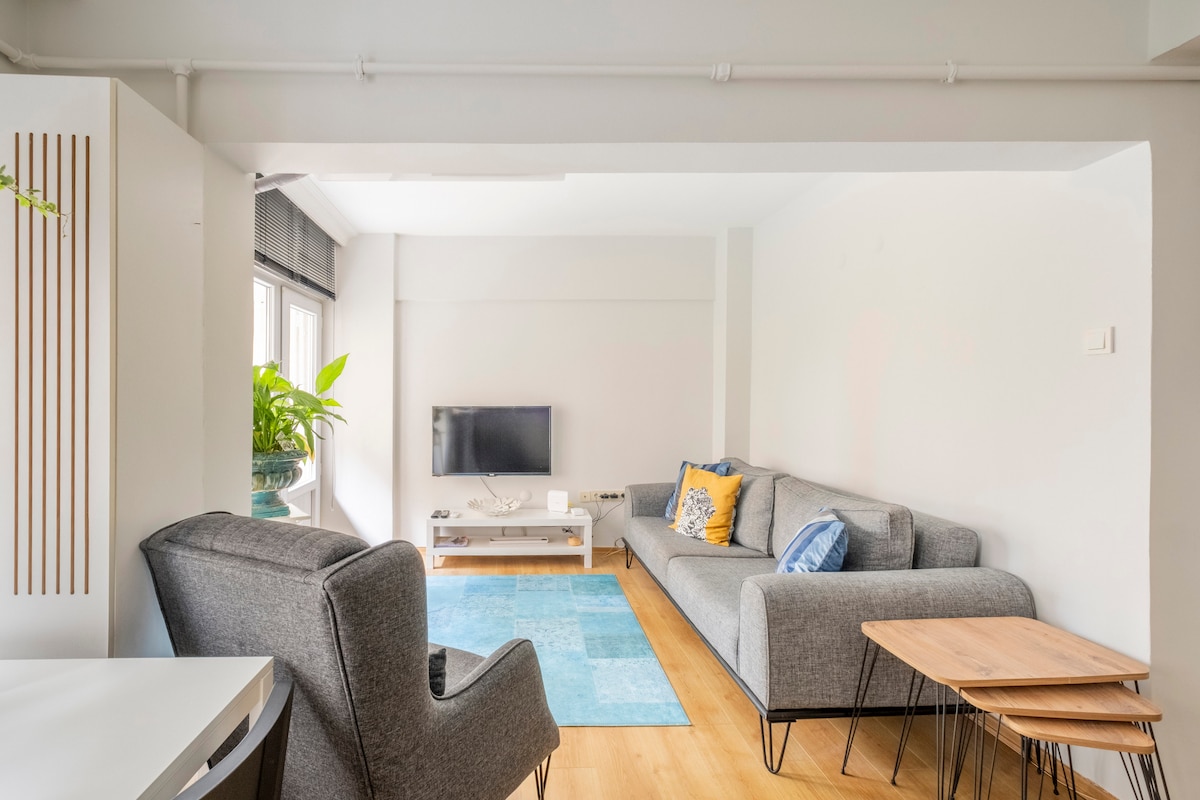 The living room features a comfortable sofa and two accent chairs arranged around a coffee table. A large television is mounted on the wall. Natural light fills the space through large windows, complemented by a vibrant blue area rug on the hardwood floor.