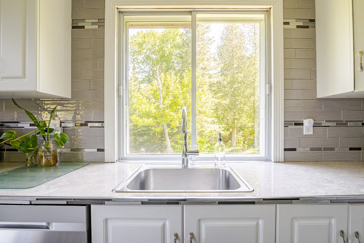A clean, well-lit kitchen is highlighted with white cabinetry and a marble countertop. A window offers a view of the greenery outside, allowing natural light to fill the space. The kitchen sink features a modern faucet, and a small potted plant adds a touch of nature indoors.