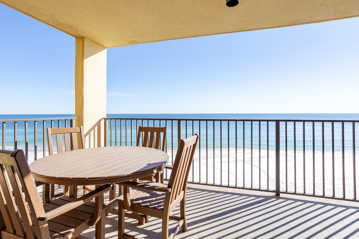 A private balcony features a round table surrounded by five wooden chairs, allowing for outdoor dining. The railing frames a stunning view of the Gulf of Mexico stretching to the horizon under a clear blue sky.