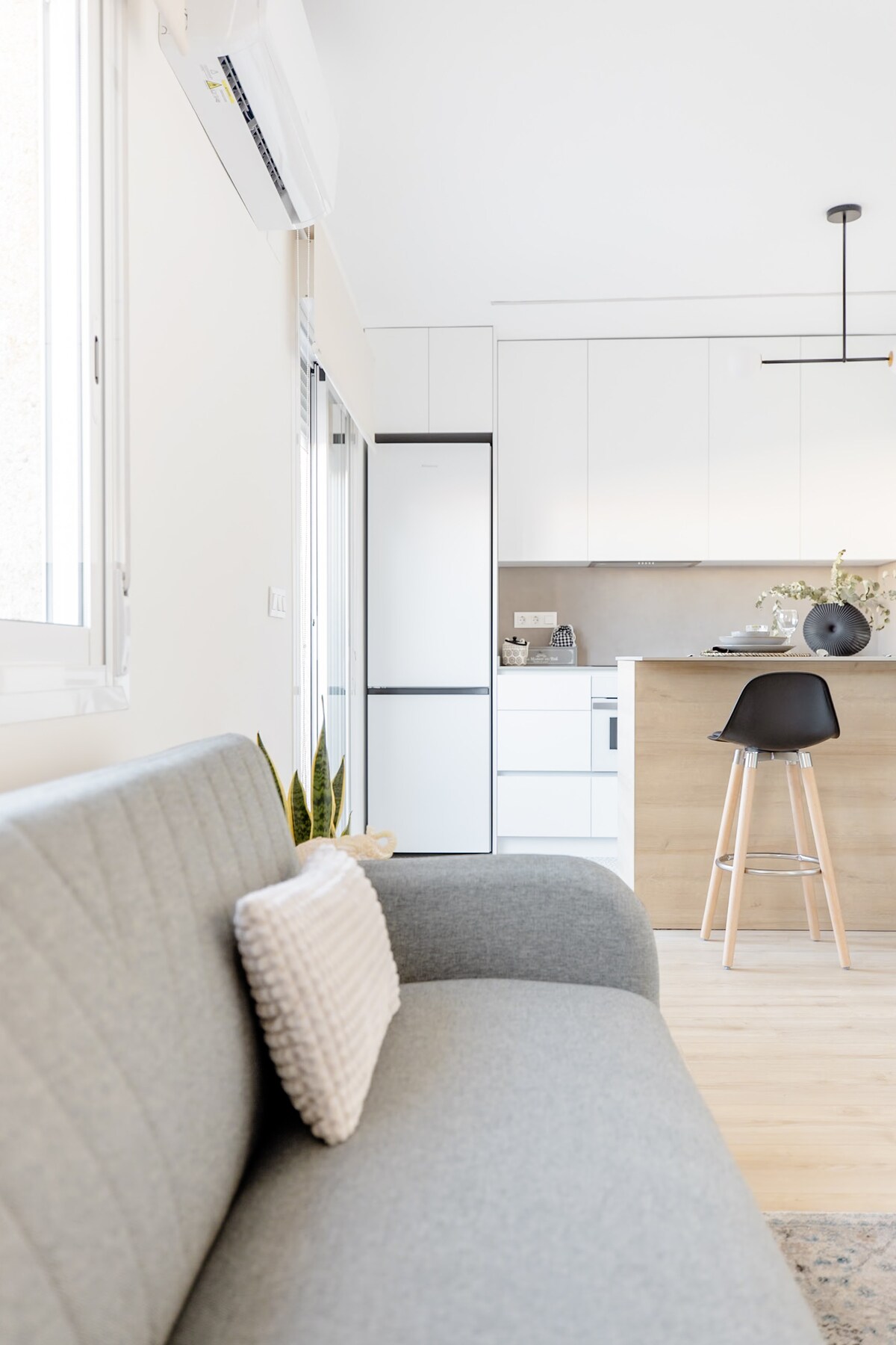 A modern living area features a grey upholstered couch with a textured pillow and a view towards a bright, open kitchen. The kitchen includes sleek cabinets and a minimalist bar with a tall stool, illuminated by a contemporary pendant light.